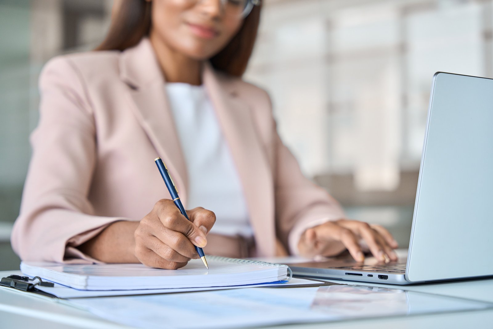 A person taking notes and working on their laptop at a desk